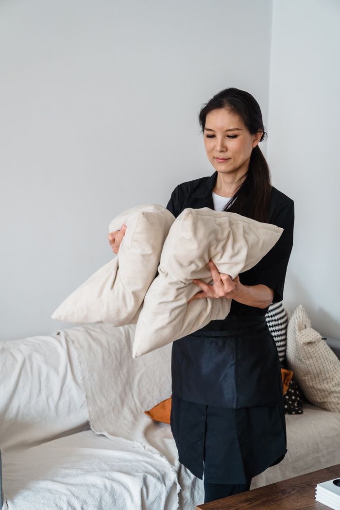Smiling housekeeper arranging pillows on a sofa in a bright living room.
